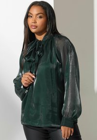 Blusa de satén verde oscuro con cuello atado, mangas largas y botones en los puños. Textura suave y brillante con un corte holgado.
