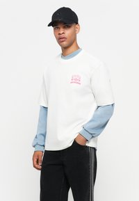 Jeune homme portant une casquette noire, une chemise blanche à manches courtes par-dessus une chemise à manches longues bleu clair, et un pantalon noir avec des rayures latérales grises, se tenant devant un fond blanc.
