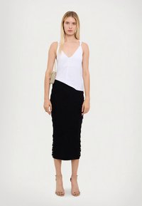 Vit ärmlös topp med V-ringning, kombinerad med en svart knälång kjol med snörningsdetaljer på sidan. Modellen bär ljusbeige klackar.