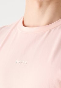 T-shirt rose clair à col rond avec petit texte blanc "BOSS" imprimé sur la poitrine, porté par une personne à la peau claire.