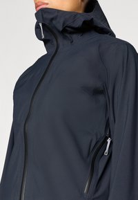 Námornícka vetrovka s vysokým golierom, zipsom vpredu a bočnými vreckami s kontrastnými kovovými prvkami a hladkou textúrou.