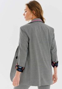 Sako s vzorom houndstooth v čierno-bielej farbe, so farebným vzorovaným podšívkou na rukávoch a ružovým prešitím na golieri.