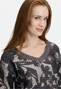 Jonge vrouw met lang rood haar die een zwart-wit gebloemde gebreide trui draagt, met een lichte glimlach, tegen een effen lichte achtergrond.