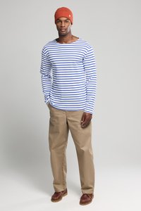 Langärmliges weißes Hemd mit blauen Streifen, kombiniert mit beige Hosen und braunen Schuhen. Besitzt eine rote Strickmütze mit Logodetail.