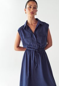 Donna che indossa un vestito senza maniche di colore navy con tasche anteriori abbottonate e vita regolabile con cordino, in piedi di fronte a uno sfondo bianco semplice.