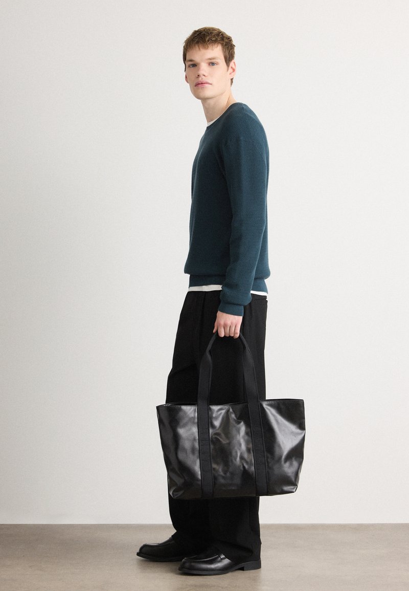 Black tote bag with glossy finish, sturdy fabric handles, and rectangular shape. Model holds it while wearing a dark sweater and black pants.
