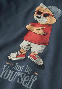 Orso dei cartoni animati con occhiali da sole rossi, maglietta rossa, pantaloncini bianchi, scarpe da ginnastica rosse e berretto grigio, con le braccia incrociate sopra il testo "Sii semplicemente te stesso."