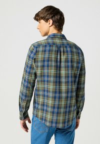 Giovane che guarda verso l'onda, indossando una camicia a maniche lunghe a quadri verdi e blu e jeans blu, su uno sfondo chiaro e semplice.