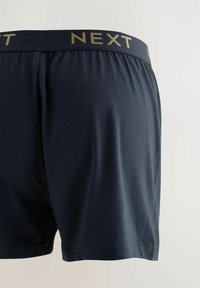 Dunkelblaue Boxershorts mit weicher Textur, versehen mit einem schwarzen Taillenbund, der mit dem Markennamen "NEXT" in goldener Schrift geprägt ist.