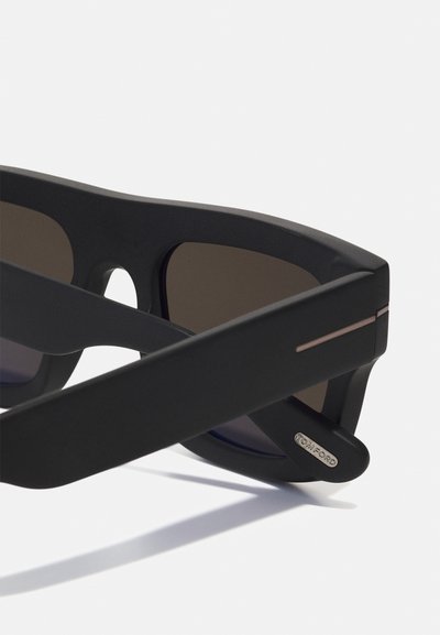 Lunettes de soleil noires avec finition mate, monture rectangulaire, verres foncés et accents métalliques subtils sur les branches. Logo gravé sur l'extrémité de la branche gauche.