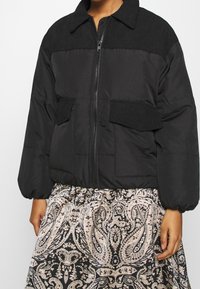 Femme portant une veste matelassée noire avec fermeture éclair et poches à rabat, associée à une jupe noire et beige à motif cachemire.