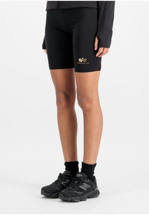 Person iført svarte tettsittende shorts med gul Alpha Industries-logo på venstre lår, svarte sokker og svarte New Balance-sneakers på hvit bakgrunn.