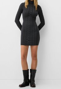 Robe gris tricotée en maille torsadée avec un col montant et des manches longues, motif texturé et silhouette ajustée, associée à des bottes en cuir noir mi-mollet.