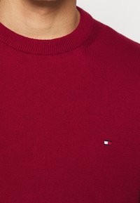 Maglione rosso a maglia con colletto tondo a coste e una piccola etichetta con logo nei colori blu, bianco e rosso, posizionata sul petto sinistro.