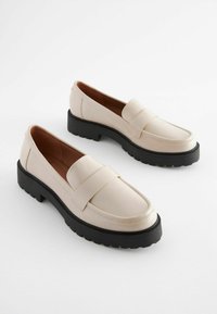 Mocasines de cuero beige con una suela de goma negra gruesa, que cuentan con una cinta ancha en el empeine y un acabado suave.