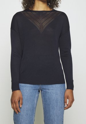 Femme portant un pull noir à manches longues avec un motif en V tricoté sur la poitrine et un jean bleu, debout les bras détendus.