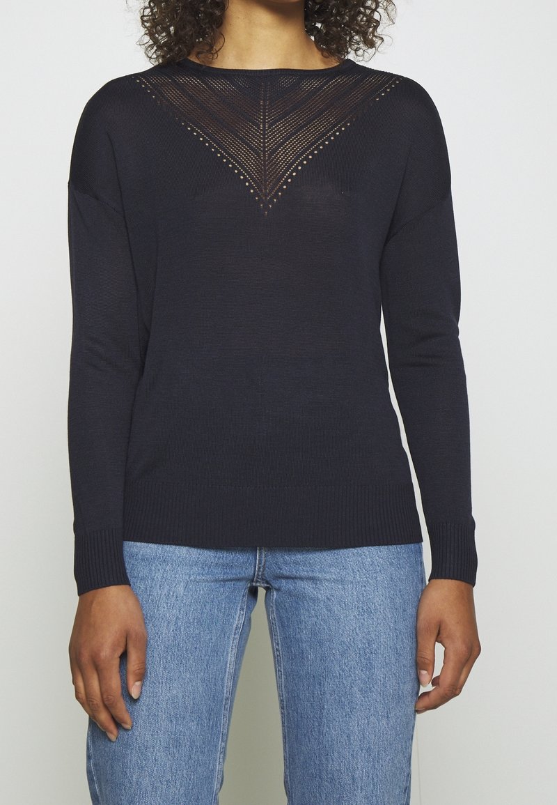 Femme portant un pull noir à manches longues avec un motif en V tricoté sur la poitrine et un jean bleu, debout les bras détendus.