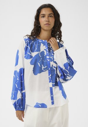 Žena s kučeravými vlasmi, oblečená v bielej blúzke s veľkými modrými kvetinovými vzormi a dlhými nafúknutými rukávmi, stojaca na jednoduchom pozadí.