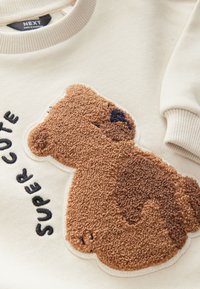 Kermainen collegepaita, jossa on ruskea pehmeä karhupäällinen, jossa on teksturoitu turkki, ja mustalla kirjailulla teksti "SUPER CUTE". Resorireunukset kauluksella ja hihoissa.