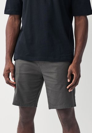 Muž oblečený v tmavosivých krátkych nohaviciach dĺžky po kolená a voľnom čiernom tričku, stojaci s uvoľnenými rukami pri bokoch.