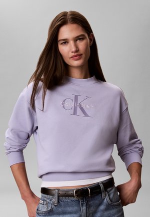 Kobieta o długich brązowych włosach, ubrana w jasnofioletową bluzę Calvin Klein i niebieskie dżinsy, stojąca na jednolitym szarym tle.