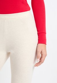Leggings de color beige claro con una textura suave y una cinturilla elástica, combinados con una blusa ajustada de manga larga roja. Primer plano de la cintura y la mano.