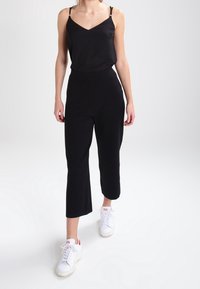 Schwarzes ärmelloses Oberteil mit dünnen Trägern, kombiniert mit schwarzen Cropped-Hosen. Das Modell trägt weiße Sneakers. Einfaches und minimalistisches Design.