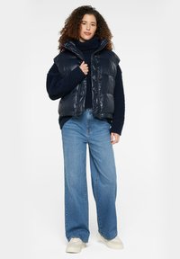 Navy puffer vest with a high collar, worn over a navy sweater. Paired with light blue wide-leg jeans and cream sneakers.