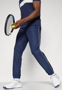 Pantaloni sportivi blu navy con polsini elasticizzati, strisce laterali bianche e una maglietta blu navy abbinata. Include una pallina da tennis e una racchetta. Sneakers bianche.