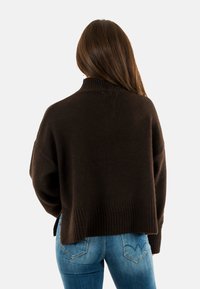 Maglione lavorato a maglia testa di moro con un alto colletto, vestibilità oversized e orlo a coste, abbinato a jeans aderenti blu chiaro.
