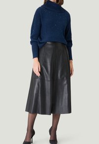 Mujer vestida con un suéter de cuello alto azul moteado, falda midi de cuero negra, medias negras transparentes y tacones altos negros, de pie contra un fondo liso.