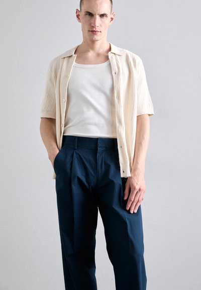 Cardigan léger à manches courtes de couleur crème clair avec texture côtelée, porté sur un débardeur blanc. Pantalons plissés bleu marine foncé. Design minimaliste et couleurs neutres.