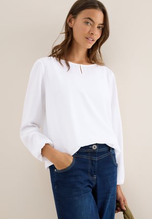 Blouse blanche à manches longues avec un col rond et un petit détail en forme de trou à l'avant, assortie à un jean en denim bleu foncé avec des poches avant.
