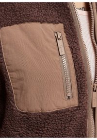 Giacca in pile marrone con una tasca in tessuto marrone e dettaglio con zip sulla parte anteriore. La texture è morbida, in contrasto con il materiale più liscio della tasca.