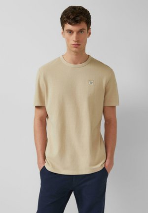 Junger Mann trägt ein beigefarbenes T-Shirt mit kurzen Ärmeln und kleinem Brustabzeichen sowie dunkelblaue Hose, steht mit den Händen in den Taschen.