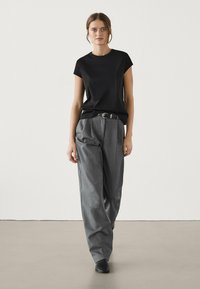 Žena kráčející vpřed, oblečená v černém krátkém rukávu, šedých širokých kalhotách s páskem a v černých špičatých botách na pozadí bez vzoru.