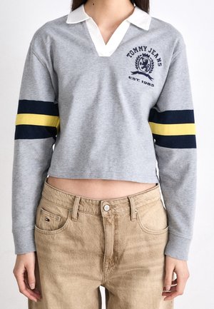 Vrouw draagt een grijze Tommy Jeans cropped top met lange mouwen, een witte kraag en gele en marineblauwe strepen op de mouwen, gecombineerd met beige broek.