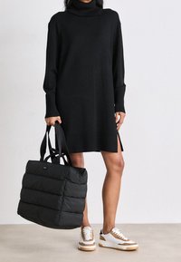 Žena v černém roláku a bílých teniskách, držící velkou černou polstrovanou tašku po svém boku.