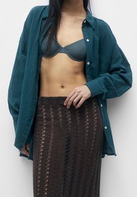 Chemise oversize bleu sarcelle en tissu léger, déboutonnée sur un soutien-gorge gris, associée à une jupe en tricot marron avec une texture à motifs.