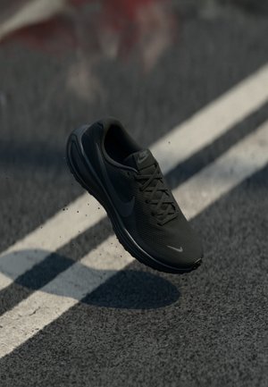 Chaussure de course noire Nike en l'air au-dessus d'une route asphaltée avec des marquages blancs et de petits débris sous la semelle.