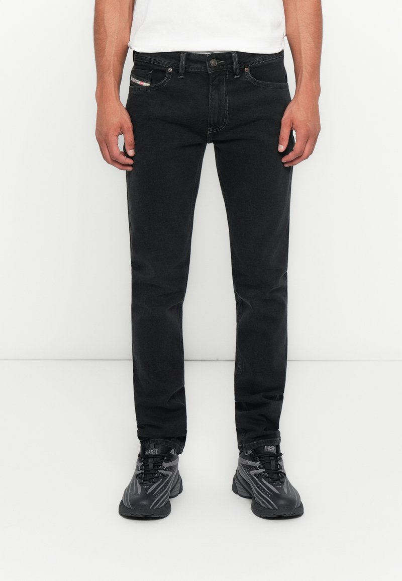 Mann, der enge schwarze Jeans, ein weißes Hemd und schwarze Sneakers trägt, steht vor einem schlichten weißen Hintergrund.