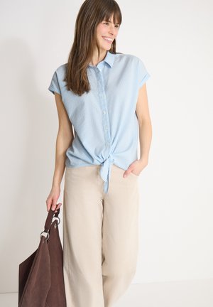 Femme souriante portant une chemise à rayures bleues claires nouée et un pantalon ample beige, tenant un grand sac à main marron foncé dans la main gauche.