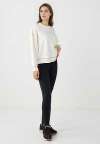 Felpa bianca con dettagli dorati, vestibilità comoda, abbinata a leggings neri e scarpe da ginnastica nere, in piedi contro uno sfondo semplice.