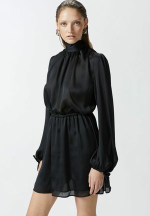 Abito mini nero realizzato in tessuto leggero e trasparente. Presenta un colletto alto, maniche lunghe a sbuffo e una vita elastica con dettagli arricciati.