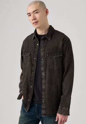 Giovane uomo con testa rasata che indossa una camicia di jeans scura lavata sopra una t-shirt nera e jeans blu su uno sfondo chiaro e semplice.