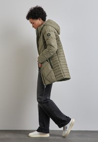 Olivově zelený, prošívaný péřový kabát s kapucí a postranními zipovými kapsami, v kombinaci s tmavými džíny a bílými teniskami, umístěný na neutrálním pozadí.