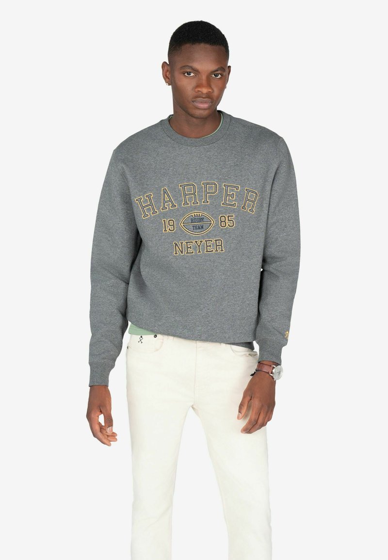 Sudadera gris con "HARPER" en amarillo, con cuello redondo y puños de canalé. Jeans blancos con un patrón sutil. Modelo de pie recto.