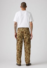 Pantalones cargo de camuflaje en beige y marrón con dos bolsillos laterales y bolsillos traseros. Combinados con una camiseta blanca lisa y sandalias negras.