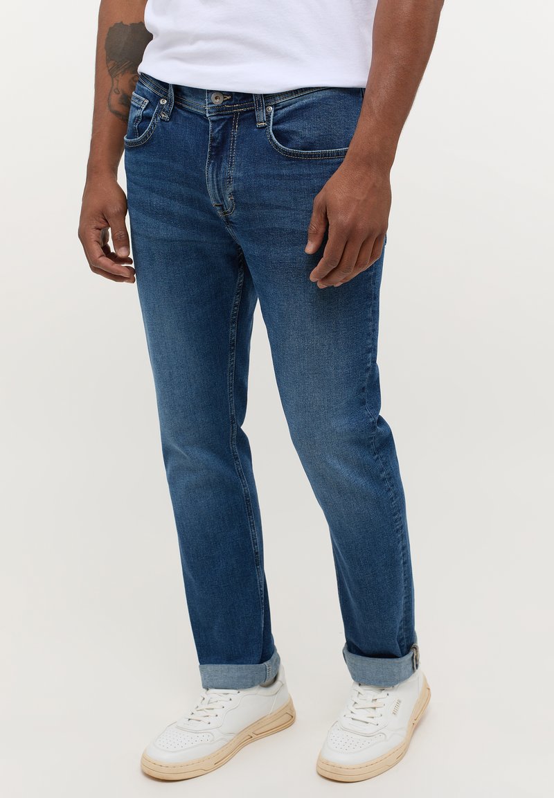 Jeans in denim blu con un taglio dritto, leggera sfumatura, cuciture visibili e orli arrotolati. Abbinati a sneakers bianche.