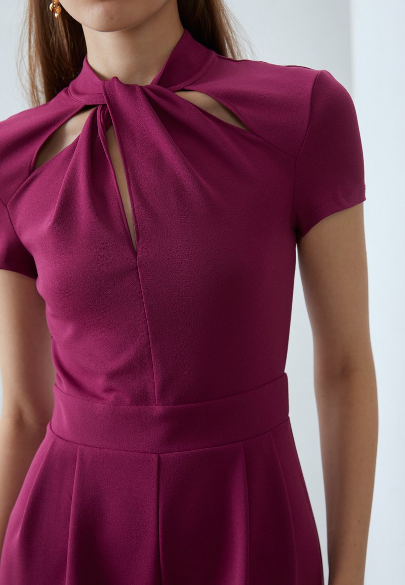 Robe ajustée magenta avec un col haut et des découpes en forme de trou de serrure, des manches courtes et une taille structurée avec des plis. Tissu doux et lisse.